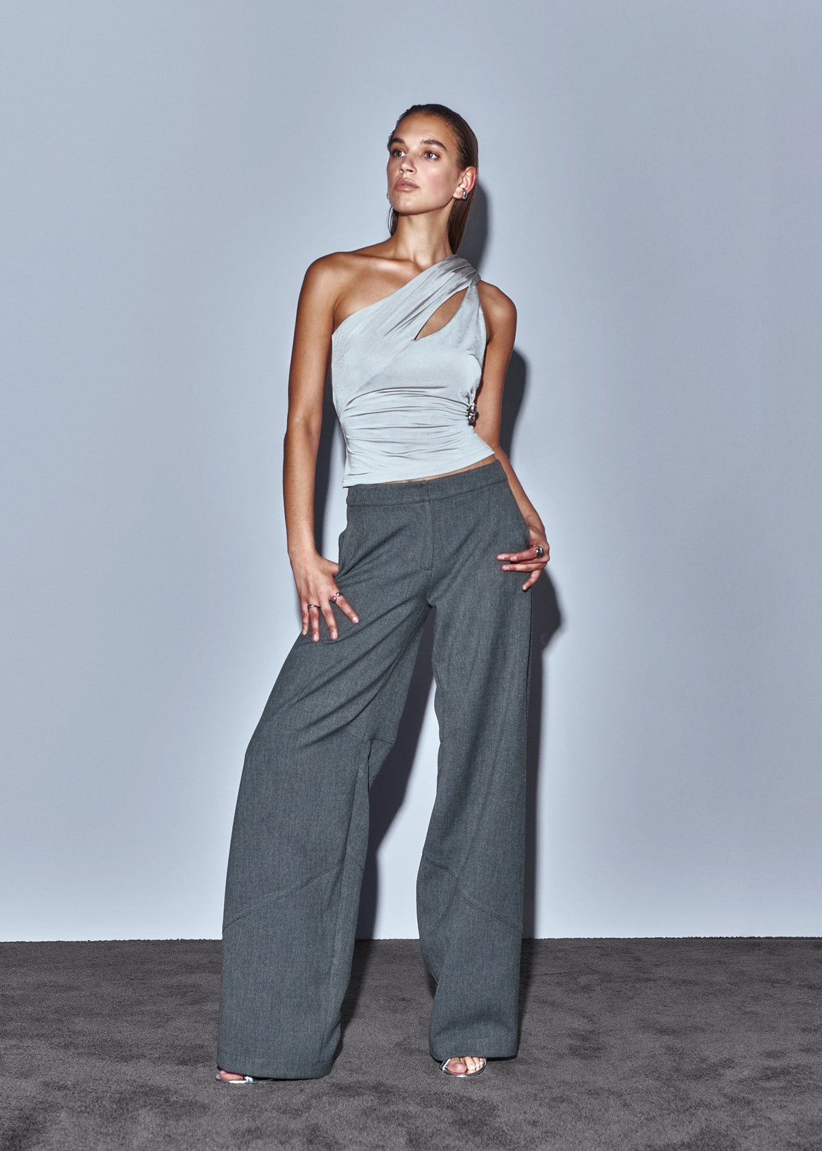 Woman wearing a light gray one-shoulder top and dark gray wide-leg pants against a plain background