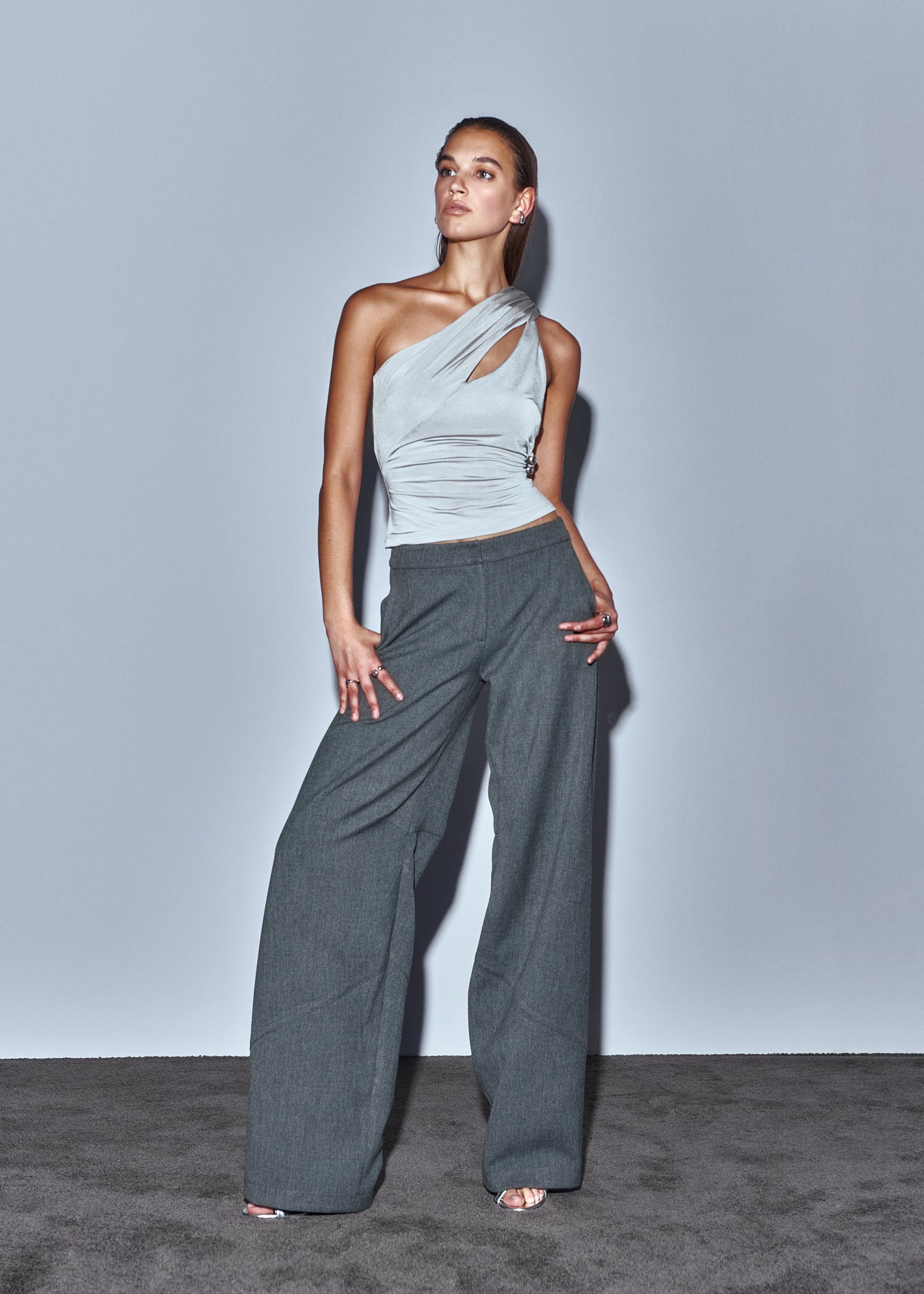 Woman wearing a light gray one-shoulder top and dark gray wide-leg pants against a plain background